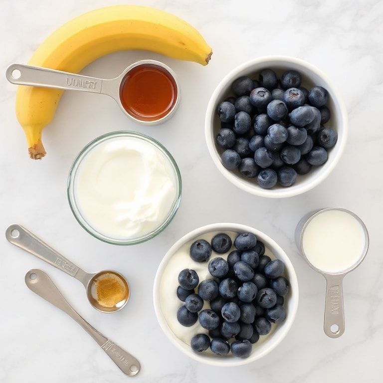 Flat lay of fresh blueberries, Greek yogurt, banana, honey, and milk in bowls, measuring cups and spoons, bright natural light over marble.
