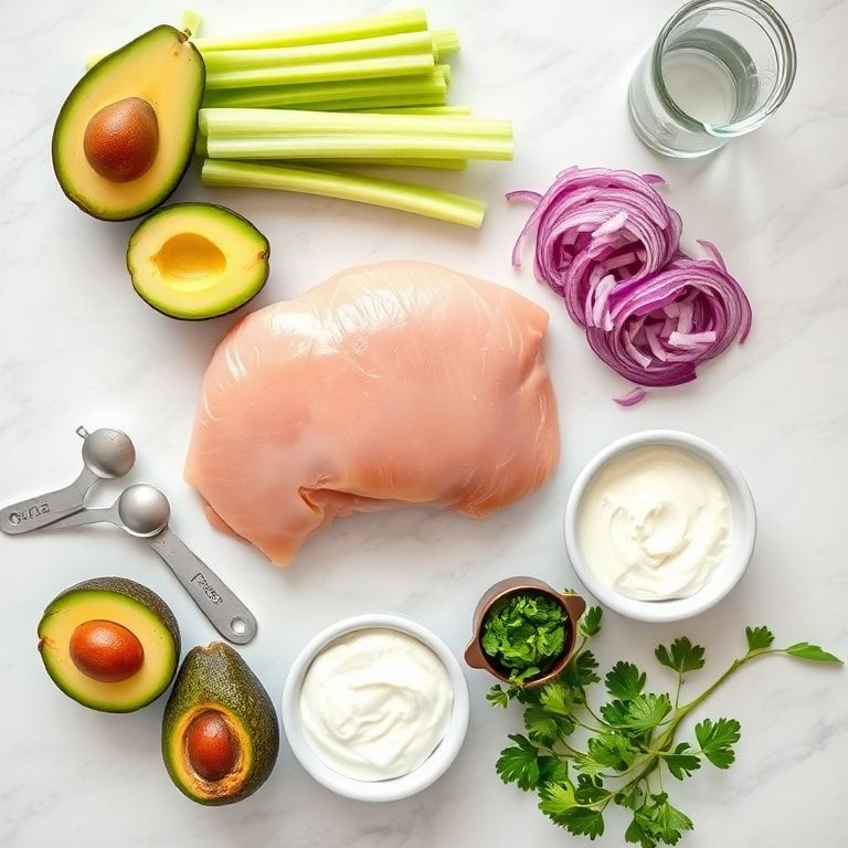 Overhead flat lay of raw chicken breast, ripe avocados, celery, Greek yogurt, red onion, lemon, and fresh parsley arranged with prep bowls and measuring spoons on white marble.